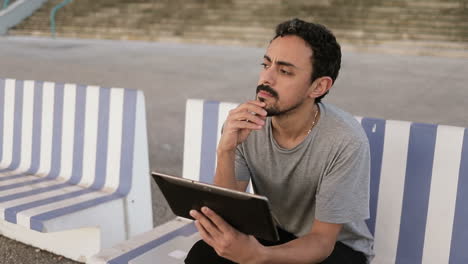 young arabic man with dark curly hair and beard in grey t-shirt sitting on quay on striped bench