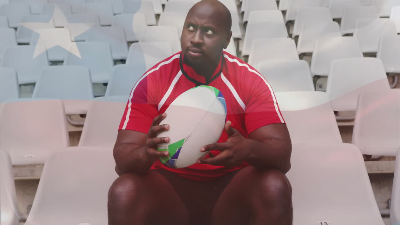 Animation of flag of chile over african american male rugby player sitting at stadium holding ball