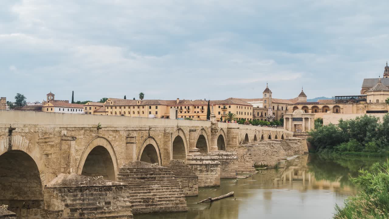 Imposing roman bridge spanning guadalquivir river in cordoba, revealing atmospheric light shifts under moody cloudscape during daylight hours