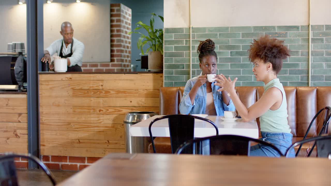 Amiche afroamericane che iniziano una conversazione, con quella in top verde che gesticola e quella in top blu che sorseggia una tazza al caffè.