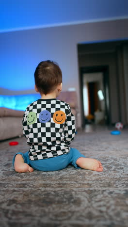 rear view of a toddler boy sitting on the floor in the room. kid rolls the pyramid rings by the floor and follows them. vertical video.