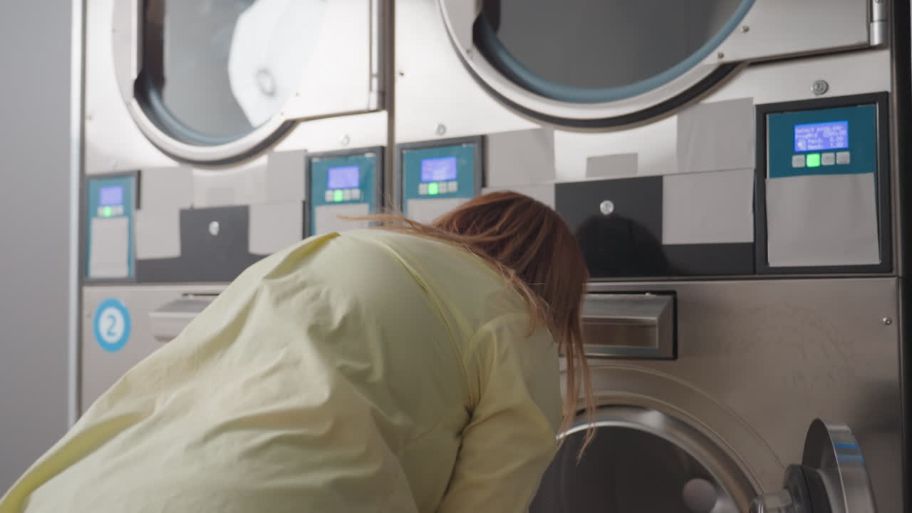 Back view employer addresses washer interface, sets basket down, loads clothes into drum inside laundromat, stainless machines in row, hygiene service workflow, indoor operation, routine chore