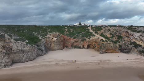 Long-aerial-shot-flying-backwards-from-a-coastline-of-rugged-cliffs-underneath-a-dramatic-sky,-over-surfers-waiting-for-waves