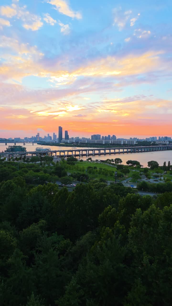 An aerial vertical pull-back shot captures the mesmerizing and colorful sunset sky over Seoul's cityscape, including the Han River, Hangang Park, and Banpo Bridge