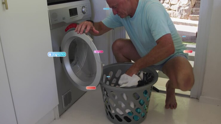 Senior man crouching at washer opening drum door loading laundry for home pulling white towels