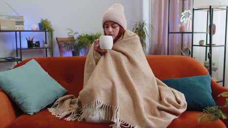 Sick-redhead-woman-wear-hat-wrapped-in-plaid-sit-alone-shivering-from-cold-on-couch-drinking-hot-tea