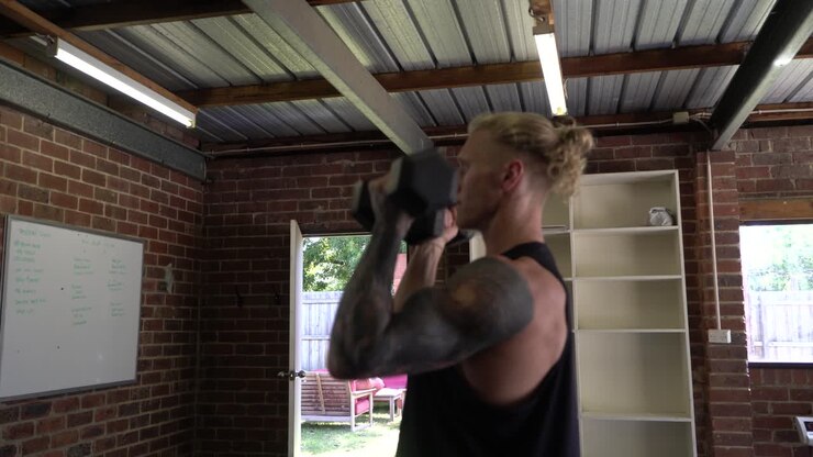 Tattoo man muscles in home gym doing dumbbell arnold press shot from side
