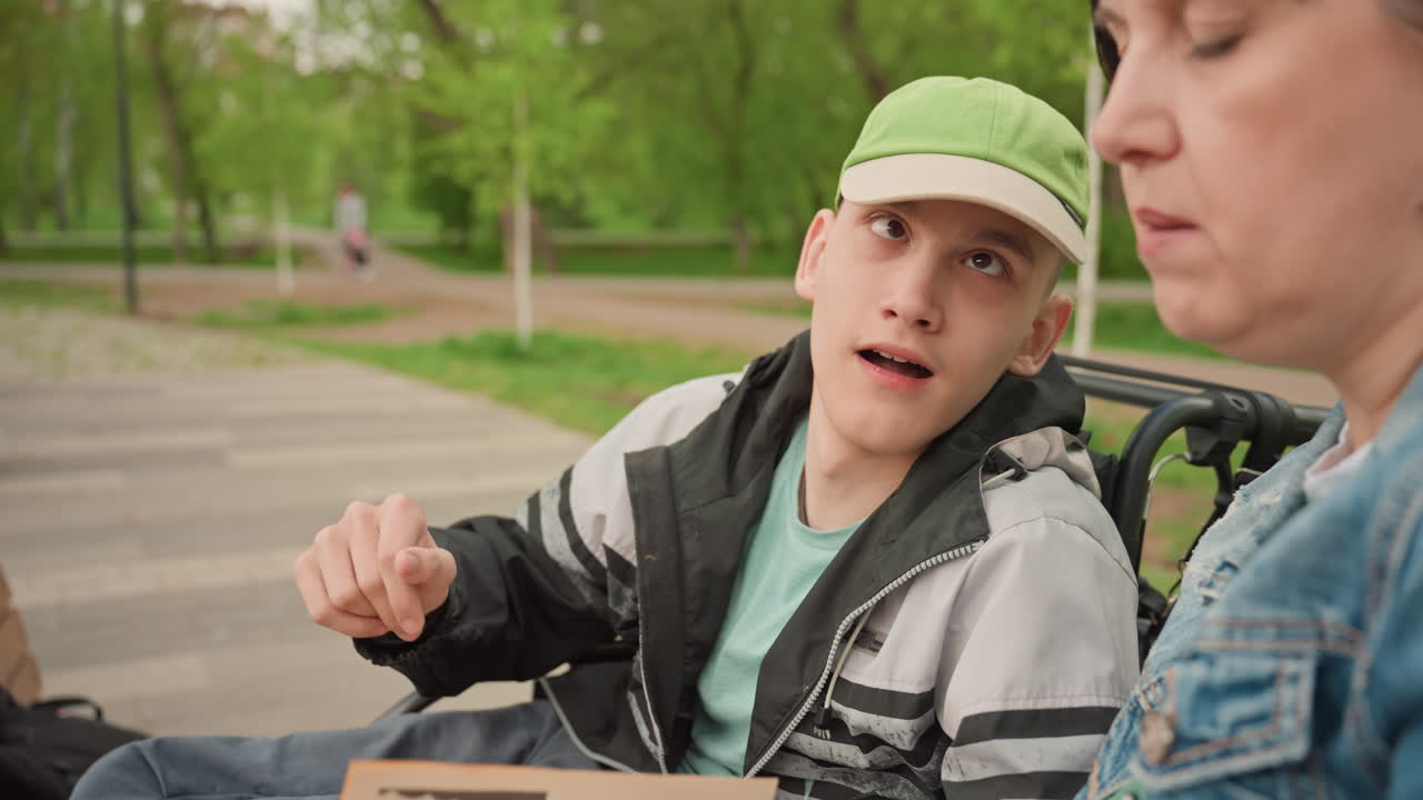 Young White Man In Wheelchair Listening While Caregiver Reads Book On Park Bench, Gentle Conversation, Leafy Spring Background, Denim Jacket, Warm Expressions, Supportive Bond, Candid Documentary Tone
