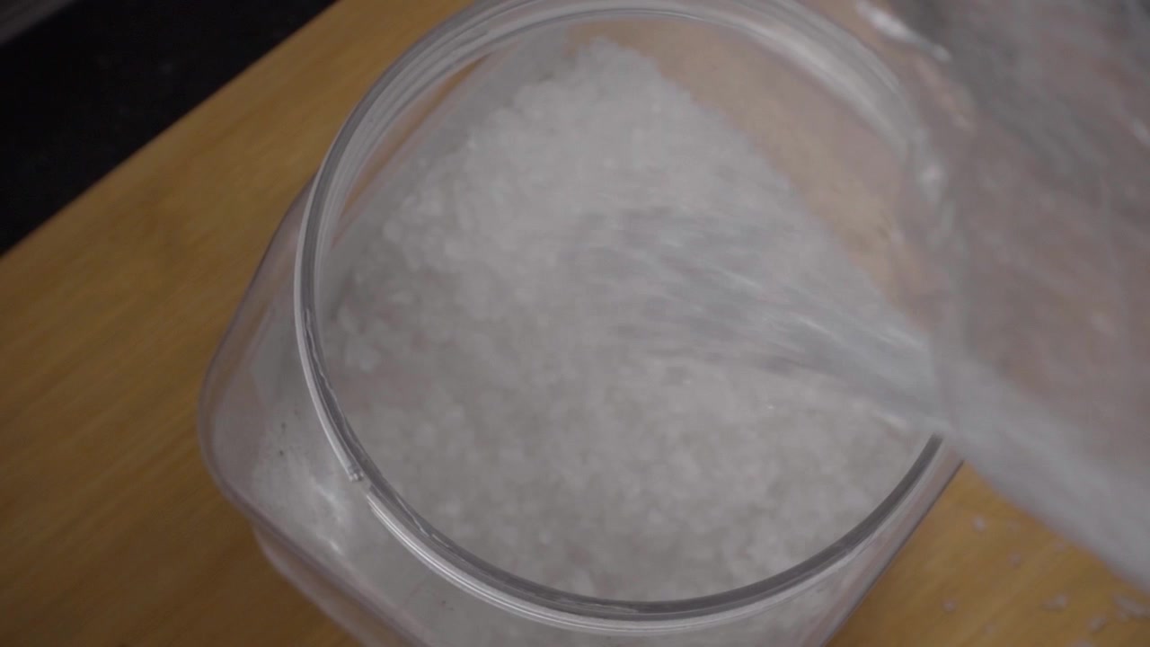 Nærbillede af granuleret sukker, der hældes i en plastikbeholder. Fine hvide sukkerkrystaller fylder opbevaringsbeholderen under madlavning i et indendørs køkkenmiljø.