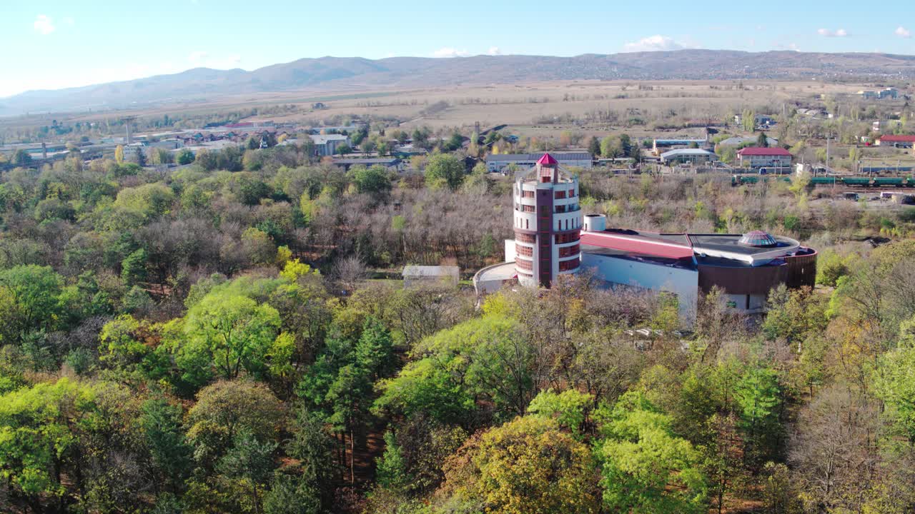 A slow-orbiting aerial shot shows a modern building with a unique spiral tower, nestled in a dense forest with beautiful autumn colors in urban Romania. Museum of natural Science, Bacau