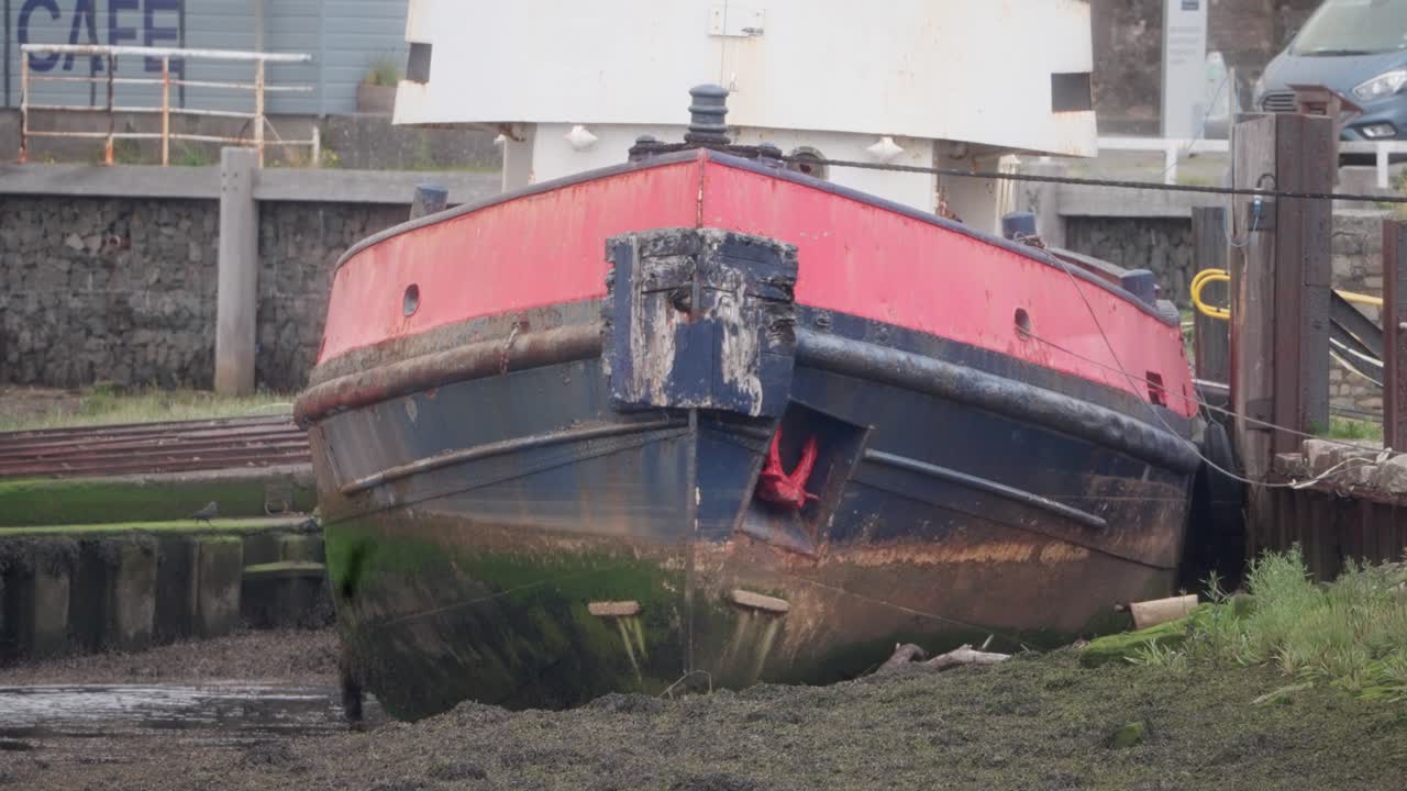 Aged red and blue barge sits grounded in mud at low tide, docked beside a seawall with visible weathering, algae stains, and marine decay. Coastal infrastructure in background