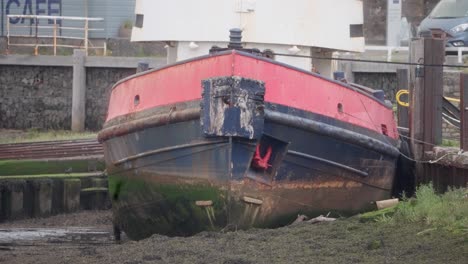 aged red and blue barge sits grounded in mud at low tide, docked beside a seawall with visible weathering, algae stains, and marine decay. coastal infrastructure in background