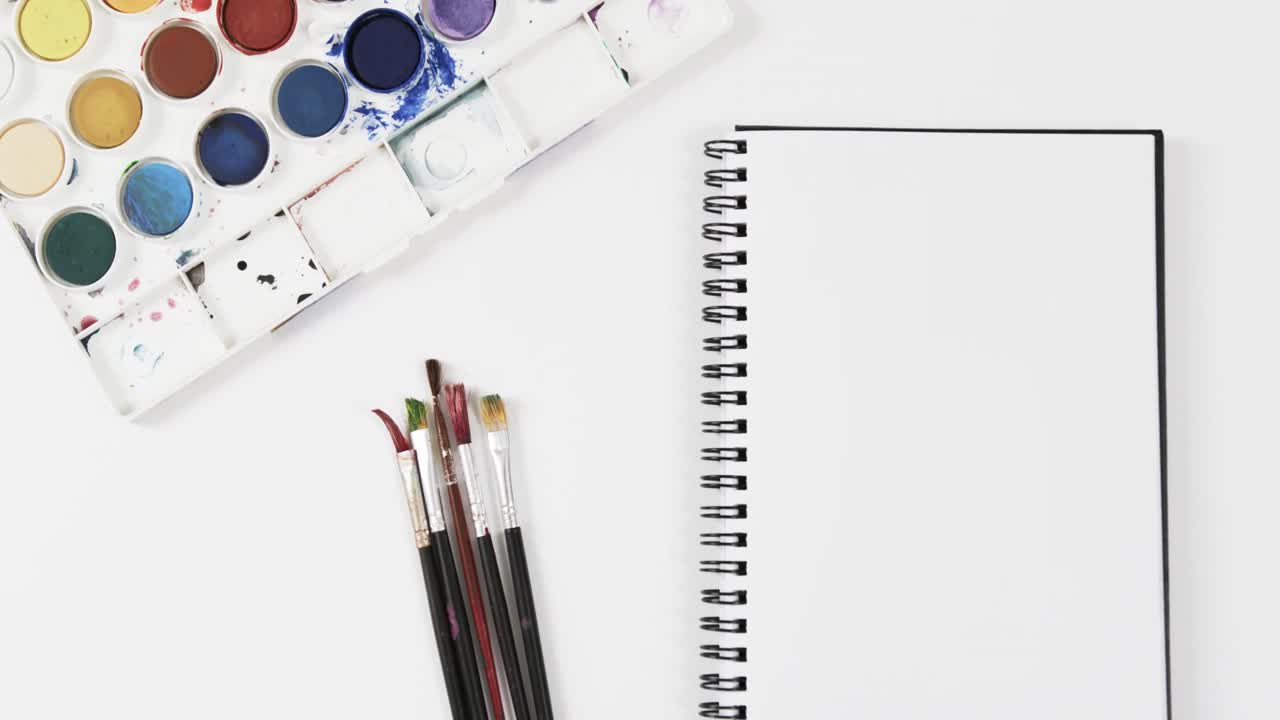 vista aérea del cuaderno, pinturas y pinceles sobre fondo blanco, en cámara lenta