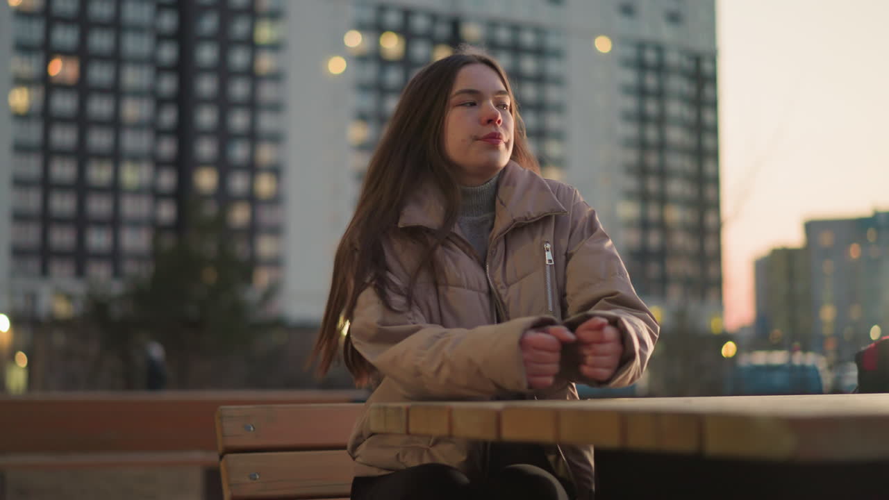A young woman dressed in a peach jacket and black trousers is seen sitting on a bench in an urban park. She places her hand on the table and gazes off into the distance with a thoughtful expression