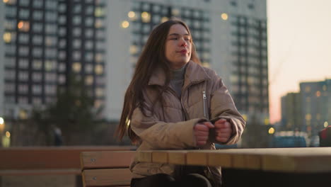 a young woman dressed in a peach jacket and black trousers is seen sitting on a bench in an urban park. she places her hand on the table and gazes off into the distance with a thoughtful expression