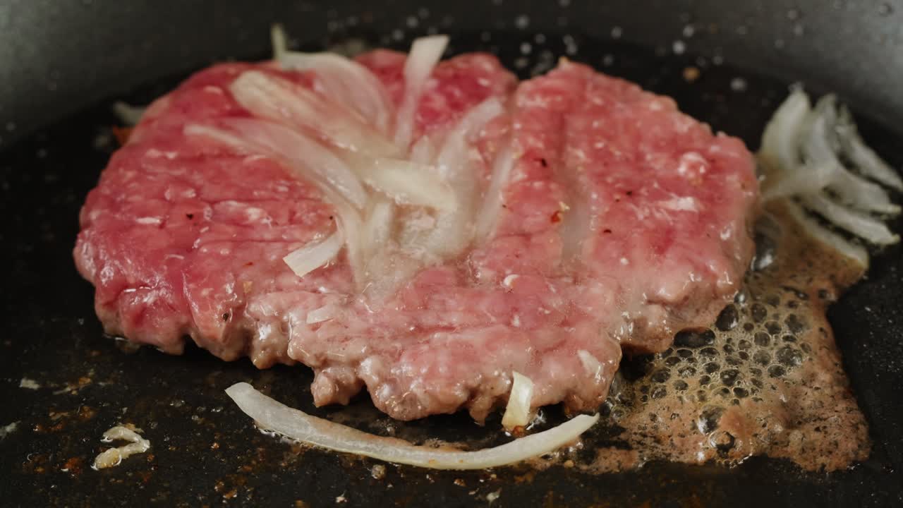 Raw hamburger patty frying in a pan with sliced onions