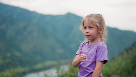 cute little girl shows thumb-up in valley against mountains