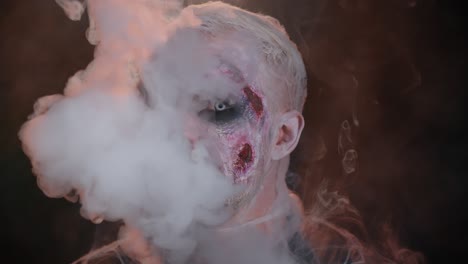 Sinister-man-horrible-scary-Halloween-zombie-looking-ominous-at-camera-blows-smoke-from-nose,-mouth