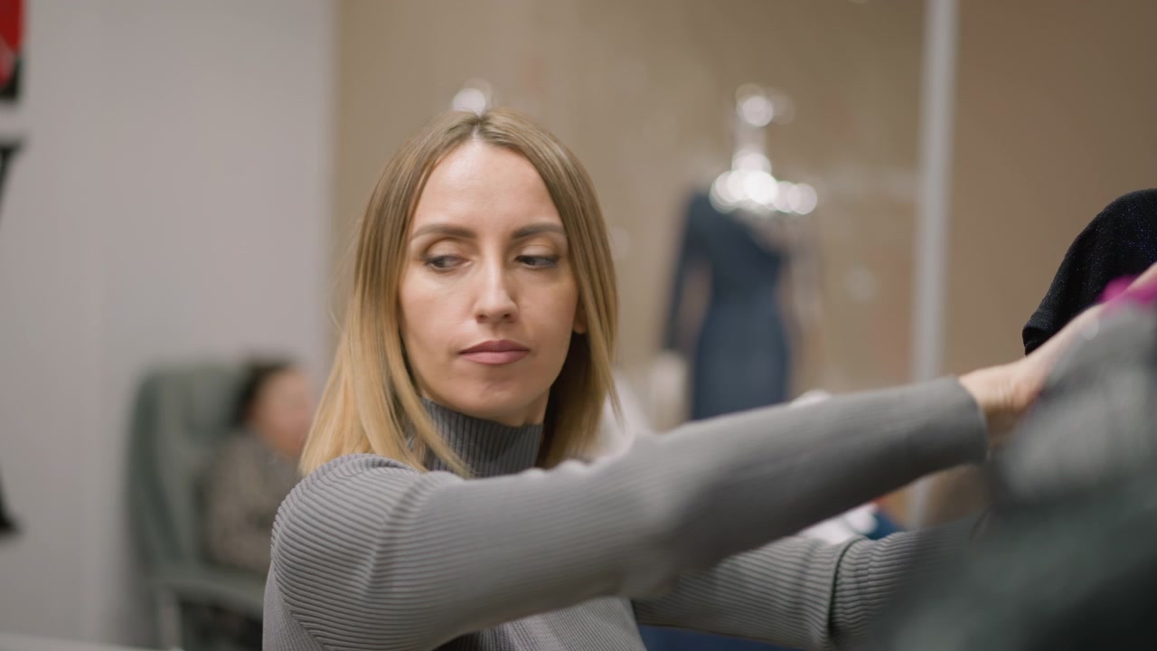 Primo piano di donna che esamina capi di abbigliamento con illuminazione interna soffusa. La donna ispeziona vari capi con manichini e altre persone visibili sfocate dietro di lei. La scena è ambientata al chiuso.