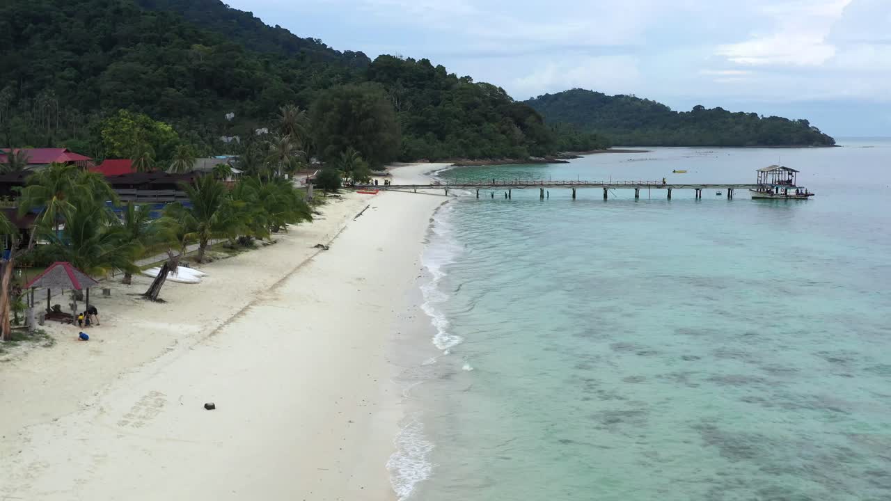 drone aéreo mover vista de playa tropical de arena blanca y agua de mar turquesa clara con pequeñas olas y palmeras bosque y puente de muelle