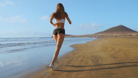 Schöne-Frau-In-Sportshorts-Und-T-Shirt-Läuft-Am-Strand-Mit-Weißem-Sand-Und-Blauem-Meerwasser-Auf-Der-Insel-In-Zeitlupe.-Wellen-Und-Sandhügel-Auf-Der-Rückseite-Haben-Gewonnen