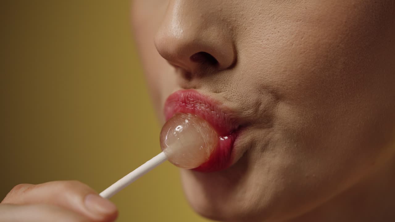 vista de cerca de una seductora mujer joven rubia con labios rojos lamiendo una deliciosa piruleta dulce y disfrutando del caramelo contra un fondo negro en cámara lenta