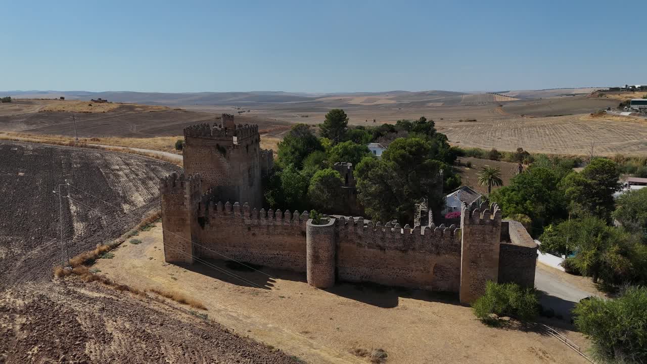 Aerial orbit of Castillo de las Aguzaderas near El Coronil, Seville, Spain, showcasing its intact stone walls and towers, set within the sunlit, dry vast Andalusian countryside