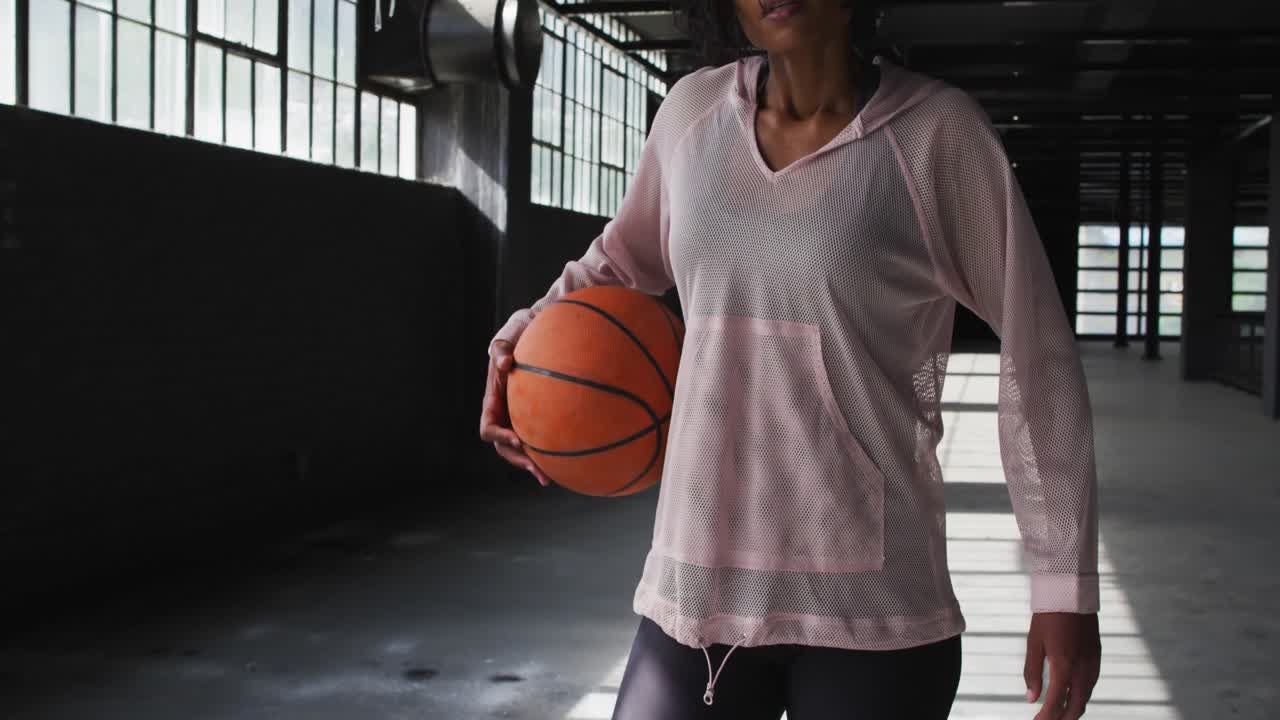 mujer afroamericana de pie en un edificio vacío jugando con un baloncesto mirando a la cámara