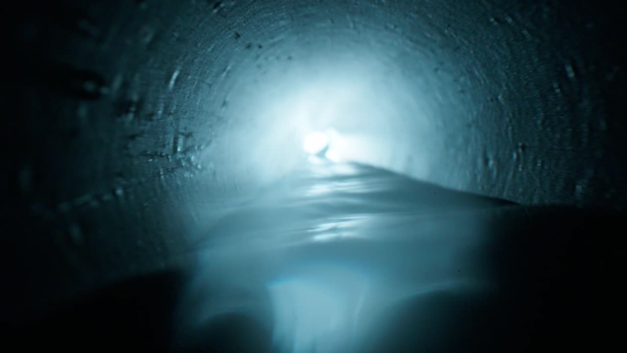 Olhando por uma longa perspectiva de um tubo de drenagem com água correndo pelo chão. A luz da extremidade distante reflete nas paredes circulares úmidas.