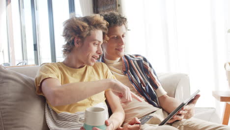 happy diverse gay male couple sitting at sofa using tablet at home, slow motion