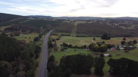 Vista-Aérea-De-Una-Carretera-Rural-Que-Atraviesa-Colinas-Verdes-Y-Una-Granja-Al-Fondo