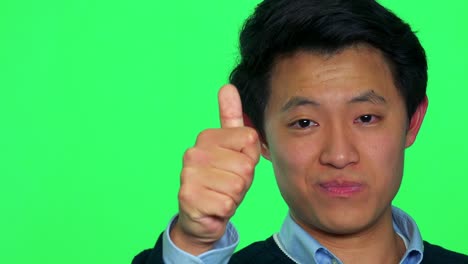 a young asian man smiles and shows a thumb up to the camera - green screen studio - closeup