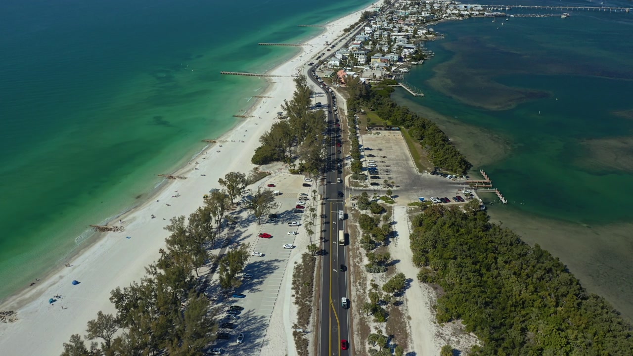 Vehicles travel along a coastal road flanked by white sandy beaches and vibrant emerald waters at Bradenton Beach. Ideal for travel and tourism content.