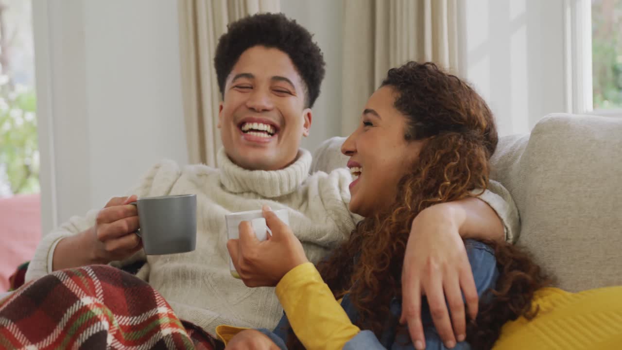 Video of happy biracial couple siting on sofa under blanket and drinking coffee