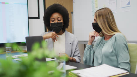 two diverse businesswomen wearing face masks working together using laptop at desk in office