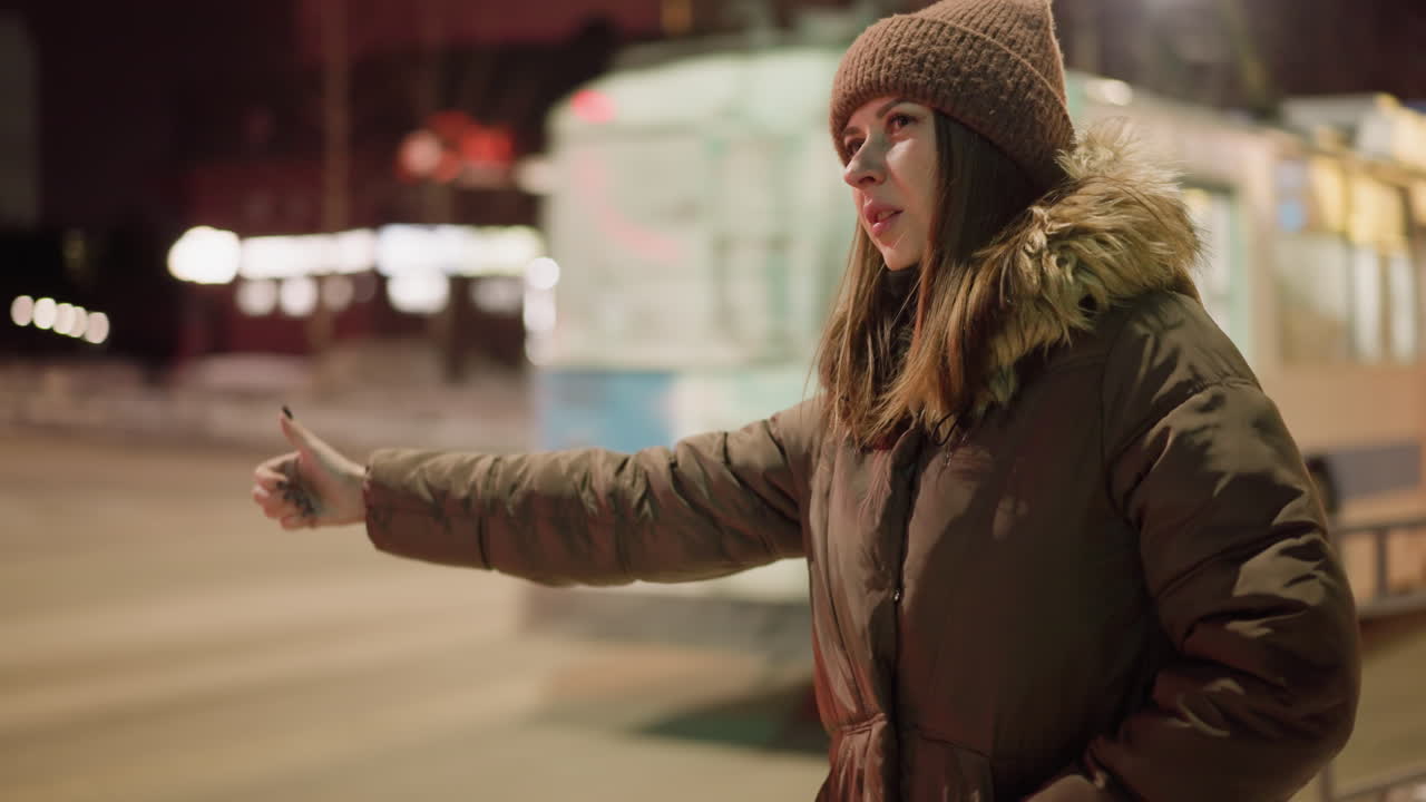 First-person view of a woman in a winter coat with a fur hood, stretching her hand out at night with buses passing by
