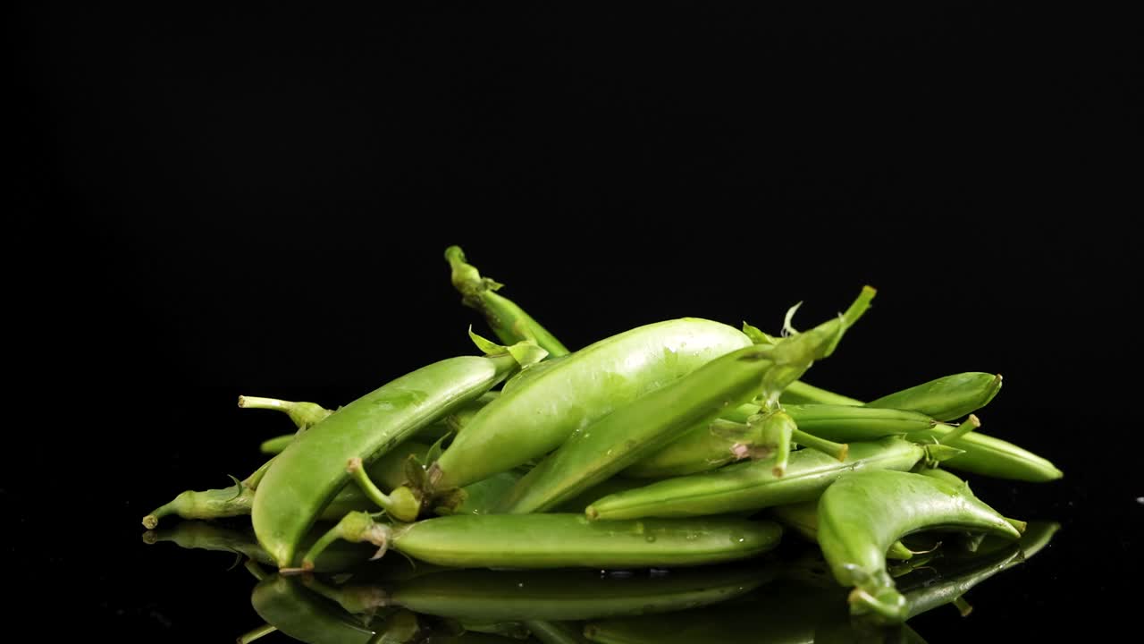 A pile of fresh snap peas rotates smoothly on a reflective black surface, highlighting their vibrant green color and texture