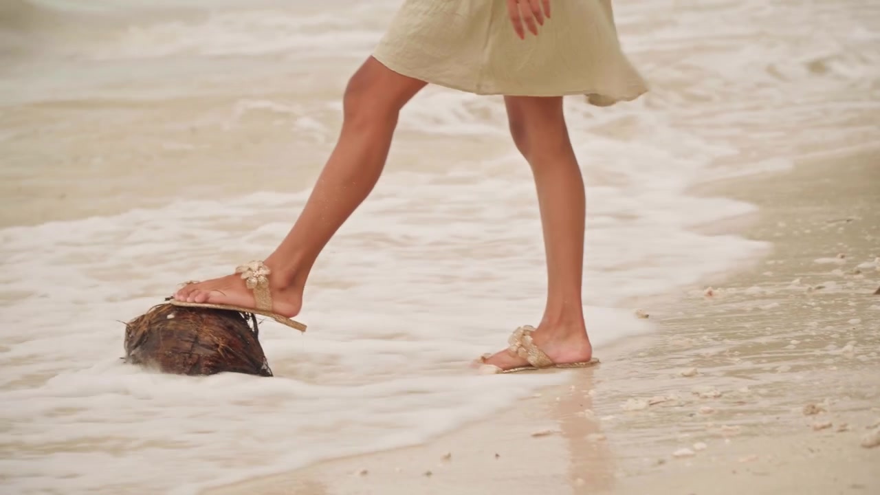 Bare feet on sandy beach shoreline. Person walking along shallow surf, gently nudging washedup coconut among shells and pebbles, waves lapping at toes, warm sunlit tones, peaceful seaside exploration