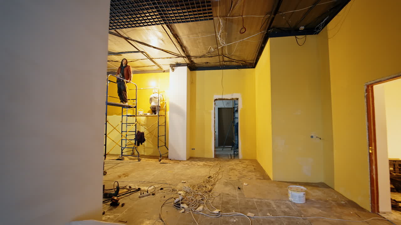 Moving from the unfinished doorway along the room being repaired. Woman paints the walls and men install ceiling light and decor.