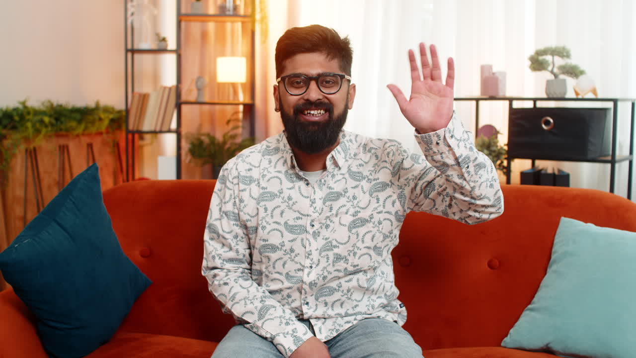 Indian man waving hand greeting while sits on sofa at home with happy expression and positive mood