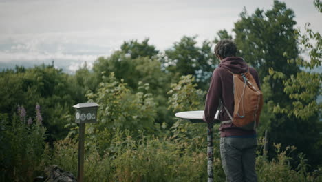 A-young-hiker-with-an-orange-backpack-is-standing-by-a-signpost-reading-and-looking-in-different-directions