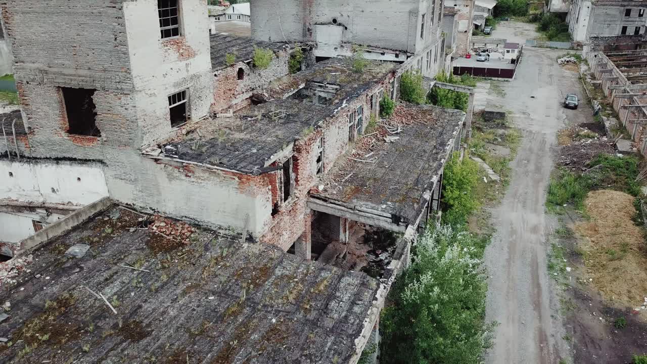 Aerial view. Ruined old building, tilt down to the dry grass and bushes. Abandoned factory halls with broken glass on windows. Apocalyptic scene with destroyed warehouse.