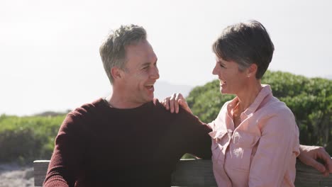 Caucasian-couple-enjoying-free-time-by-sea-on-sunny-day-sitting-on-bench-and-talking