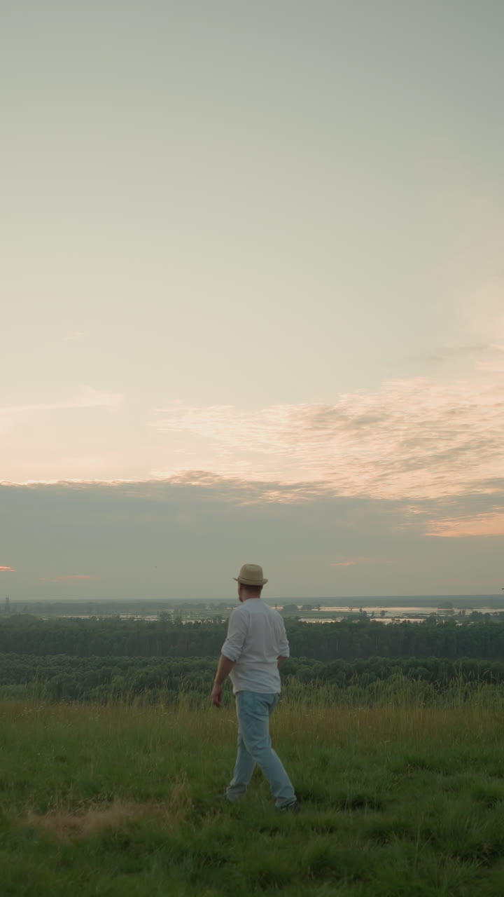 un hombre vestido con una camisa blanca, sombrero y vaqueros camina a través de un campo cubierto de hierba hacia un lago sereno al atardecer. se detiene con los brazos cruzados, contemplando el paisaje pacífico y las aguas tranquilas