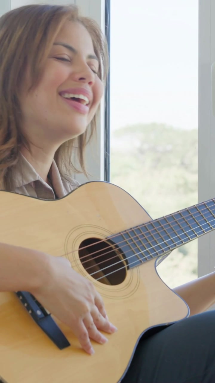 Video verticale: Una donna, indossando un top e un orologio, posiziona la chitarra e strimpella cantando a casa, vicino alla finestra.