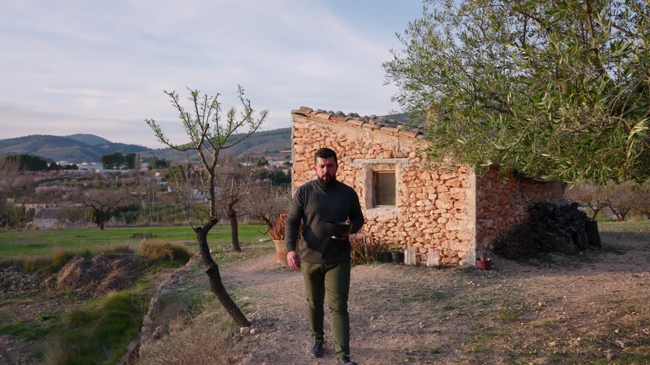 hombre camina entre los árboles de almendra en flor al atardecer, con una antigua cabaña de piedra de estilo mediterráneo a principios de primavera