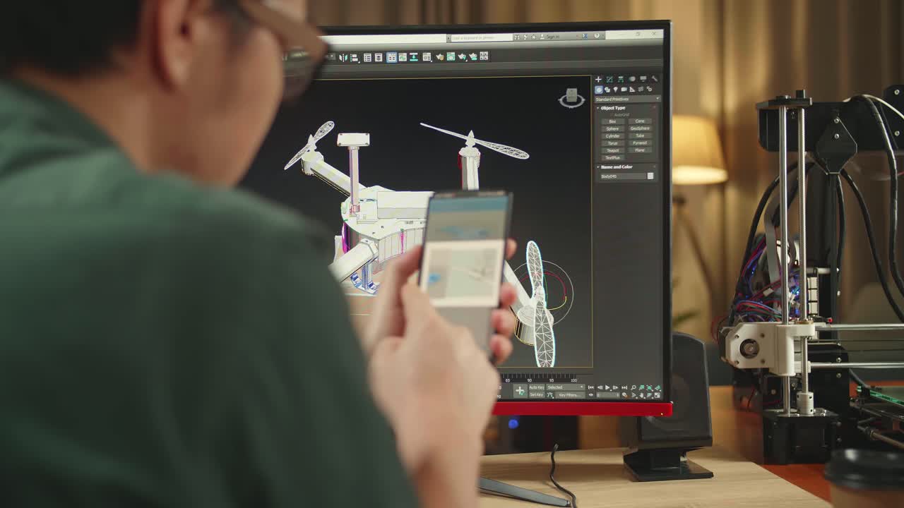 Over The Shoulder Of Asian Engineer Using Mobile Phone While Work On Personal Computer And 3D Printer, Screen Shows Cad Software With 3D Prototype Of Drone Delivery