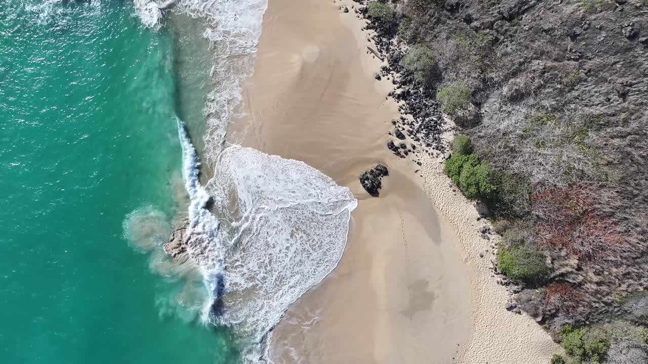 Aerial top-down drone footage of ocean waves breaking on a sandy beach in Oahu, Hawaii, showcasing turquoise water, white surf, tropical coastline, and the rhythmic motion of waves along the shore