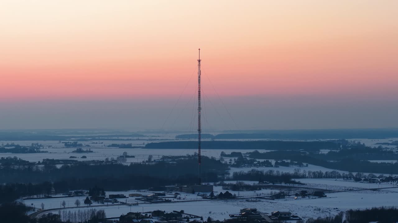 ドローン映像が捉えた、高い通信塔がそびえ立つ印象的な冬の景観。夕焼け空が温かい輝きを放ち、下の雪景色と対照的だ