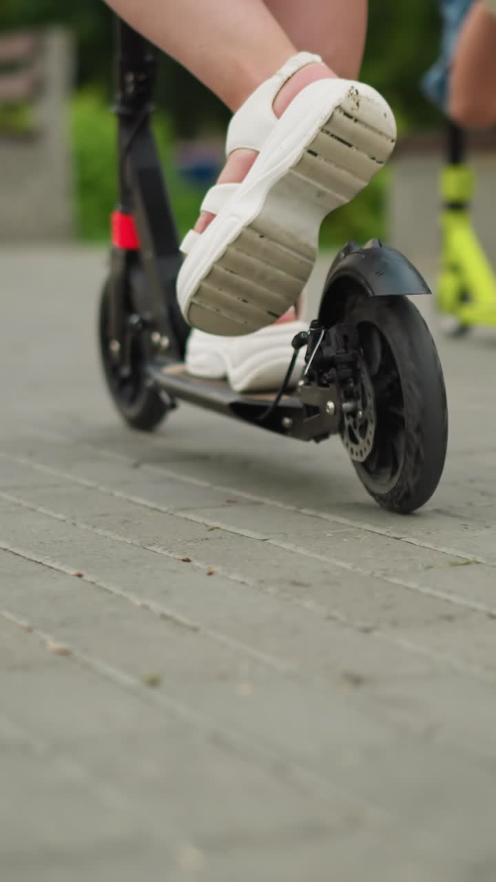Close-up of a woman riding a black scooter on a paved walkway, her legs are visible as she moves forward, with a child riding a green scooter in the background fast pass her